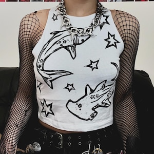 May include: A white sleeveless crop top featuring a black graphic print of a shark and a hammerhead shark surrounded by various star shapes. The top is worn with black fishnet sleeves and a thick, chunky silver-toned metal chain necklace.