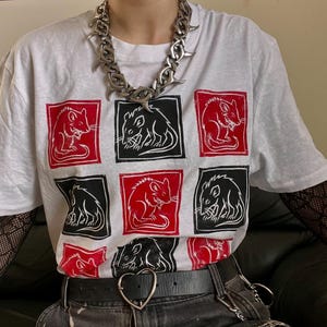 Peut inclure: Un t-shirt blanc à manches courtes orné d'une grille de neuf carrés, chacun contenant une illustration stylisée de rat en encre rouge et noire alternée. Un collier à chaîne métallique argentée épaisse aux maillons pointus repose sur l'encolure.