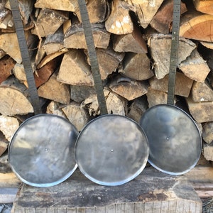 Puede incluir: Tres utensilios de cocina redondos de metal con mangos largos. Los utensilios están colgados en una pared de leña apilada.