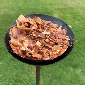 May include: A black metal smoker box filled with wood chips. The chips are various shades of brown and tan. The smoker box is on a metal pole, with a green grassy background.