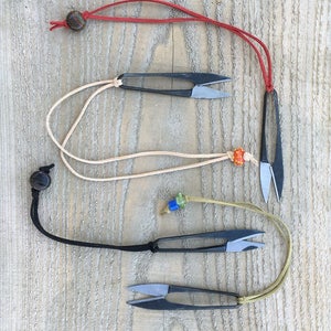 Puede incluir: Cuatro pares de pequeñas tijeras de bordado negras con cordones y cuentas de diferentes colores. Los cordones son rojo, beige, negro y verde.