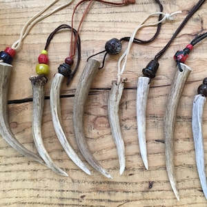 May include: A collection of seven necklaces featuring curved, light brown antler pendants suspended from thin leather cords. Each pendant is accented with decorative beads in various colors including red, yellow, black, and dark brown, attached at the top.