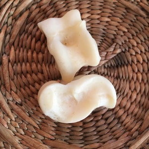 May include: Two off-white, organically shaped soaps rest on a woven, circular, brown wicker mat. The soaps have a smooth texture and a slightly irregular form, suggesting a handmade quality. The soaps are positioned to overlap slightly.