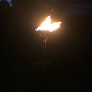 Puede incluir: Una antorcha de metal con un acabado negro y un diseño alto y delgado. La antorcha está encendida y tiene una llama naranja brillante.