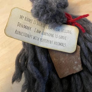 May include: A handmade gray wool doll with a tag that reads, "MY NAME IS SIGRID FROM JELLING, DENMARK. I AM LEARNING TO CARVE RUNESTONES WITH DIFFERENT ANIMALS." The tag is tied with red yarn. A small, brown stone is attached.