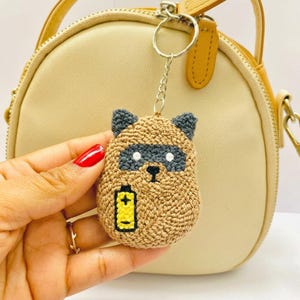 May include: A crocheted raccoon keychain with a silver keyring. The raccoon has a brown body, gray mask and ears, and white eyes. A yellow battery graphic is on the raccoon's belly. The keychain is held in front of a beige purse.