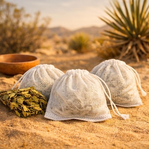 Peut inclure: Trois sacs blancs à cordon remplis d'herbes sont posés sur du sable, à côté d'un tas de feuilles vertes séchées et d'un bol en bois. Les sacs ont une texture ajourée. L'arrière-plan montre des plantes du désert et un ciel brumeux.