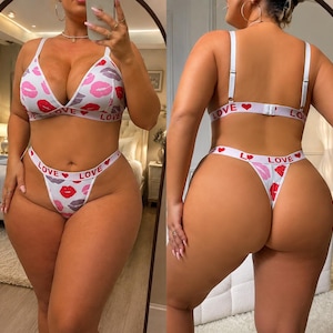 May include: A two-piece lingerie set featuring a white triangle bra and matching thong with a red and pink lip print. The elastic bands on both pieces are white with the repeating red text "LOVE" and small red heart icons throughout the design.