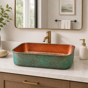 Peut inclure: Une vasque rectangulaire à la texture martelée, dotée d'un intérieur en cuivre brillant et d'un extérieur à la finition vert sarcelle patinée. Le lavabo est accompagné d'un grand robinet en laiton brossé centré derrière sur un comptoir blanc.