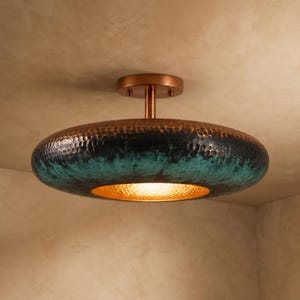 Puede incluir: Una lámpara de techo de montaje empotrado con una pantalla ancha en forma de disco y textura de metal martillado. El exterior muestra una pátina oscura oxidada con tonos azul verdoso y cobre, mientras que el interior revela un acabado dorado.