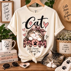 Puede incluir: Una camiseta de color crema con un gráfico de un gato atigrado sonriente que lleva una diadema rosa con estampado de leopardo. El texto dice Cat, Mom y Same Purrson Different Day, junto a corazones rosas y una huella en un corazón.