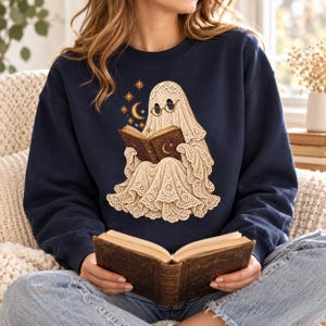 Puede incluir: Una sudadera azul marino de cuello redondo con un gráfico de un fantasma color crema que lleva gafas redondas y lee un libro. El fantasma está rodeado de pequeñas estrellas doradas y una luna creciente. El tejido del fantasma parece encaje.