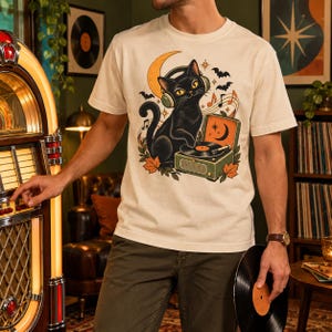 Puede incluir: Una camiseta de manga corta color crema con un gráfico de un gato negro con auriculares mientras utiliza un tocadiscos vintage. El diseño incluye una luna creciente, notas musicales, murciélagos y hojas de otoño rodeando al gato.