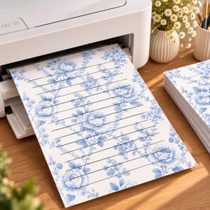 Puede incluir: Una hoja de papel de carta blanco con un patrón repetitivo de rosas azules y delicadas enredaderas florales, superpuesto con finas líneas horizontales negras para escribir. El papel sale de una impresora blanca, con una pila de papel al lado.