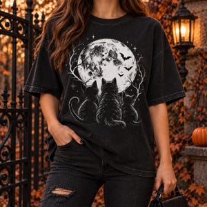 Puede incluir: Una camiseta negra de manga corta con un estampado gráfico blanco de tres gatos sentados de espaldas, mirando hacia una gran luna llena rodeada de ramas de árboles desnudas, pequeñas estrellas y varios murciélagos volando en el cielo nocturno.