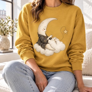 Puede incluir: Una sudadera de cuello redondo color amarillo mostaza con una ilustración gráfica de un personaje de luna creciente con una túnica oscura, sentado sobre una nube blanca y cosiendo una estrella con aguja e hilo. La sudadera tiene mangas largas y puños acanalados.