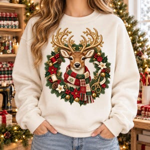 Puede incluir: Una sudadera de cuello redondo color crema con un gráfico central de un ciervo que lleva una bufanda con estampado rojo, verde y blanco. El ciervo está enmarcado por una corona navideña decorada con carretes de hilo, botones, tijeras y flores de pascua.