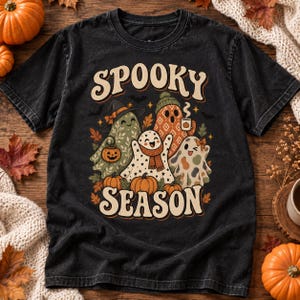 Puede incluir: Una camiseta negra de manga corta con un estampado gráfico que incluye el texto SPOOKY SEASON. El diseño muestra tres fantasmas decorados con patrones, hojas de otoño, pequeñas calabazas y murciélagos en tonos naranja, verde y crema.
