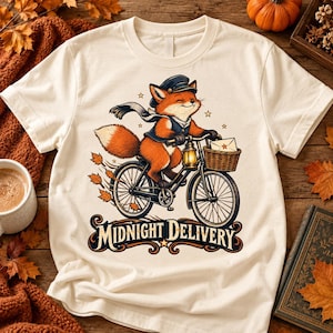Puede incluir: Una camiseta de manga corta color crema con el gráfico de un zorro con gorra y bufanda, montando una bicicleta con una linterna y una cesta que contiene un sobre. El texto MIDNIGHT DELIVERY está impreso debajo en una fuente decorativa.