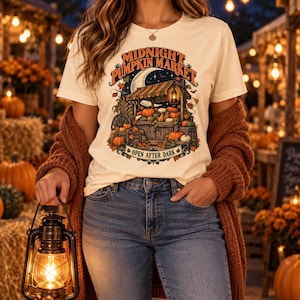 Puede incluir: Una camiseta de manga corta color crema con un gráfico de un puesto de mercado de calabazas bajo una luna creciente. El texto dice "MIDNIGHT PUMPKIN MARKET" y "OPEN AFTER DARK" con ilustraciones de calabazas, hojas y una linterna.