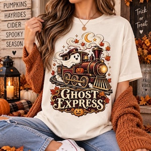 Puede incluir: Una camiseta de color crema con un gráfico de un tren de vapor conducido por un fantasma, con el texto "GHOST EXPRESS" debajo. El diseño incluye hojas de otoño, una luna creciente y calabazas en tonos naranja, marrón y amarillo.