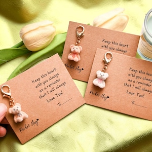 May include: A close-up of three small, light brown cards with a teddy bear charm attached. Each card has the text "Keep this heart with you always as a reminder that I will always Love You!" and "Pocket Hug."