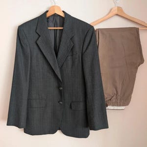Op de afbeelding: Een donkergrijze krijtstreep blazer met twee knopen en een revers met inkeping hangt aan een houten kleerhanger. Daarnaast wordt een bruinbeige broek ook op een houten kleerhanger getoond. De kledingstukken hangen tegen een lichtroze muur.
