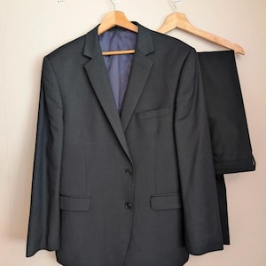 Peut inclure: Un costume gris anthracite composé d'une veste et d'un pantalon assorti, suspendus à des cintres en bois. La veste a deux boutons et deux poches avant. Le costume est présenté sur un mur de couleur neutre.