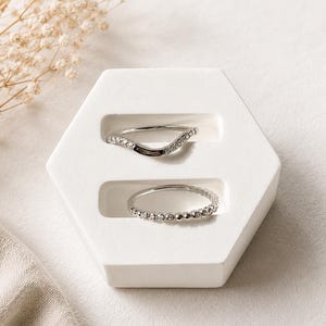 Può includere: Due anelli color argento su un supporto esagonale bianco. L'anello superiore presenta una fascia curva a forma di onda incastonata con piccole pietre chiare. L'anello inferiore è una fascia circolare sottile con una trama a perline.