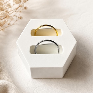 Può includere: Un espositore per anelli esagonale bianco con due fessure orizzontali. Una fessura contiene un sottile anello in metallo color oro, mentre l'altra contiene un sottile anello in metallo color argento. Il supporto ha una finitura opaca liscia e geometrica.
