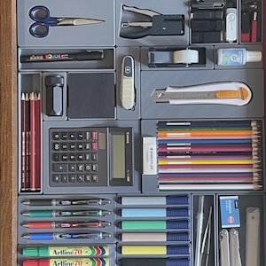 May include: A grey plastic desk drawer organiser containing various office supplies including a metal ruler, blue scissors, a stapler, a black marker, a pocket knife, a utility knife, a calculator, coloured pencils, pens, markers, and a multi-tool.