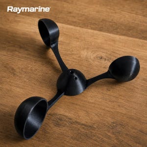 Può includere: Un anemometro nero con tre bracci a forma di coppa che si estendono da un mozzo centrale. La parola "Raymarine" è stampata in bianco nell'angolo in alto a sinistra. Il dispositivo è appoggiato su una superficie di legno.
