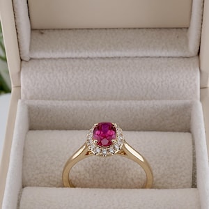 May include: A gold ring featuring an oval-cut vibrant pink gemstone surrounded by a halo of small white stones. The ring is presented inside a beige jewelry box with the text Hello Diamond printed in gold on the inside of the lid.
