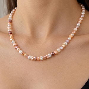 May include: A necklace composed of small, irregularly shaped pearls in varying shades of white, cream, peach, and soft purple. The pearls are strung together in a single strand, creating a textured, multi-colored appearance around the neck.