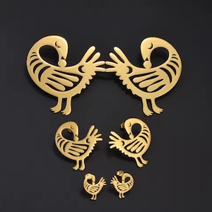 May include: Gold-colored Adinkra bird jewelry set. The set includes two large pendants, two medium pendants, and two small stud earrings. The bird design features a curved neck, a beak, and a stylized body with intricate details. The jewelry is displayed on a black background.