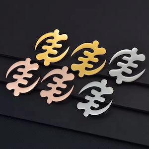 Op de afbeelding: Zes Adinkra-symbolen in goud, roségoud en zilver zijn gerangschikt op een donkere ondergrond. De symbolen hebben een gebogen, abstract ontwerp, met een centraal element en een halvemaanvorm aan één kant.