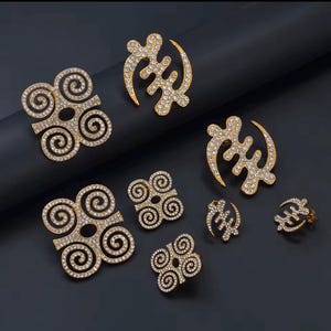 May include: A selection of gold-coloured jewellery items, including earrings, rings, and pendants, decorated with sparkling clear crystals. The designs feature Adinkra symbols, set against a dark backdrop.