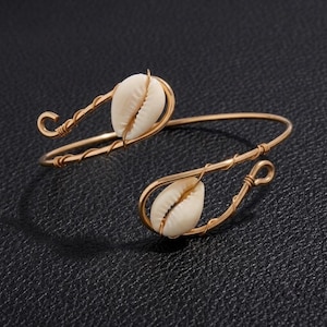 May include: A gold-toned wire cuff bracelet featuring two white cowrie shells. The wire is wrapped around the base of each shell, and the ends of the cuff are formed into small loops. The design is open-ended, allowing for adjustment to the wrist.