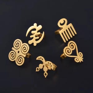 May include: Five gold-colored rings with various Adinkra symbols. The rings are displayed on a dark background. The symbols include a comb, a heart shape, a spiral design, and other abstract shapes. The rings are made of a shiny metal.