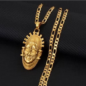 May include: A gold-toned necklace featuring a figaro chain and a pendant shaped like a stylized human face mask. The pendant has a textured surface with a beaded border and radiating spikes along the top edge, showcasing a polished metallic finish.