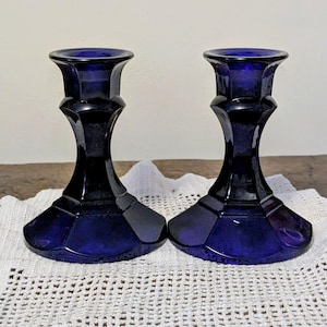 May include: Two cobalt blue glass candle holders. Each holder has a wide, geometric base, a slender, faceted stem, and a flared top. The candle holders are displayed on a white, lace-like doily.