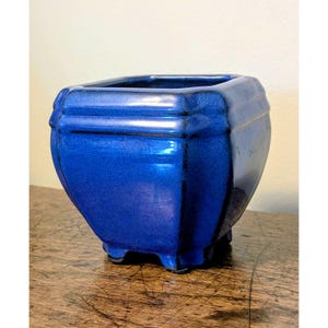 Può includere: Un vaso quadrato in ceramica con una finitura lucida color blu cobalto intenso. Il contenitore presenta un bordo a gradini, angoli arrotondati e quattro piccoli piedini integrati alla base, che offrono una solida stabilità a questo oggetto.