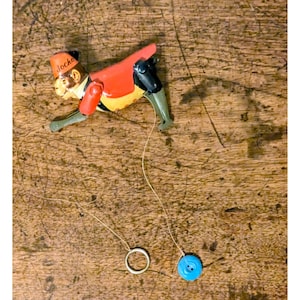 Könnte beinhalten: Ein Vintage-Zugspielzeug mit einem Affen, der als Jockey verkleidet ist. Der Affe trägt eine rote Jacke, eine schwarze Hose und einen Fez mit dem Wort "Jocko". Das Spielzeug hat einen gelben und grünen Körper mit einem blauen Knopf und einem silbernen Ring.