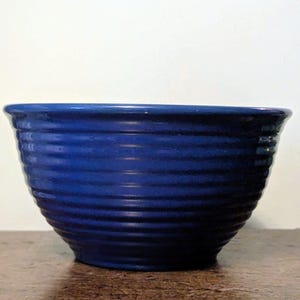 May include: A deep blue ceramic mixing bowl featuring a series of horizontal, raised ridges encircling the exterior. The bowl has a wide, flared rim and a smooth, glossy finish that reflects light across its curved, circular body.