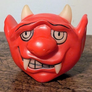 Peut inclure: Une figurine en plastique rouge représentant une tête de diable avec deux cornes blanc cassé, de grandes oreilles pointues et un large sourire malicieux. Le visage présente des yeux cernés de noir, un nez proéminent et deux crocs visibles.
