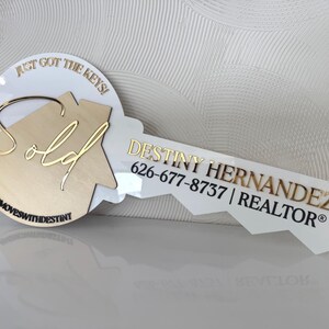 May include: A key-shaped sign with the words "Just Got The Keys!" and "Sold" in gold lettering. The sign also includes the name "Destiny Hernandez, Realtor" and a phone number. The sign is white with a gold and wood-tone design.