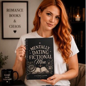 Op de afbeelding: Een zwart spiraalnotitieboek met de witte tekst MENTALLY DATING FICTIONAL Men. IT'S COMPLICATED. en illustraties van een open boek en rozen. Het notitieboek wordt rechtop gehouden en toont een omslagontwerp met sterren en florale accenten.