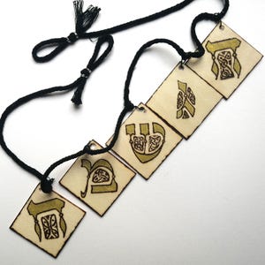 Puede incluir: Un collar de cordón negro con cinco colgantes cuadrados de madera. Cada cuadrado de madera clara está decorado con un diseño oscuro e intrincado de nudos de inspiración celta en tinta dorada. El cordón incluye pequeñas borlas negras.