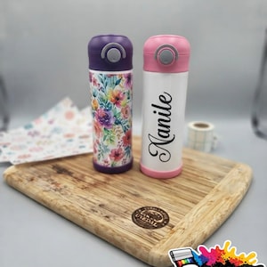 Puede incluir: Dos botellas de agua cilíndricas con tapas abatibles. La botella de la izquierda presenta un patrón floral colorido con flores moradas, rosas y naranjas. La botella de la derecha es blanca con el nombre Nanile en negro y una base rosa.