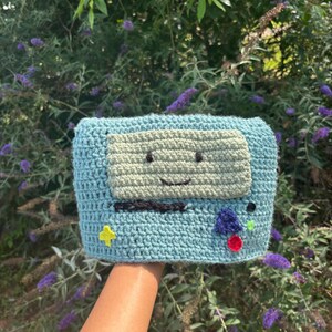 May include: A light blue crochet pouch featuring a rectangular light green face with embroidered black eyes and a mouth. The front includes a black horizontal line, a yellow cross, a small purple triangle, a red circle, and a tiny green dot.